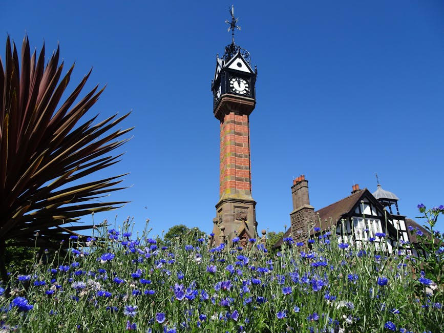 A tall, ornate clock tower constructed from red brick with decorative stone detailing at its base, standing prominently in a small garden. The tower features a classic clock face with black hands and
