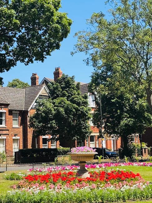 A tall, ornate clock tower constructed from red brick with decorative stone detailing at its base, standing prominently in a small garden. The tower features a classic clock face with black hands and